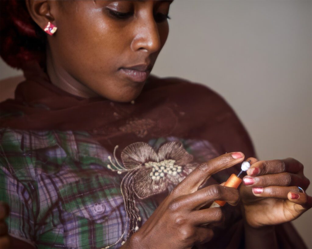 Ethiopian woman in plaid shirt concentrating on detailed handicraft work, holding small beaded item in her hands with painted nails.